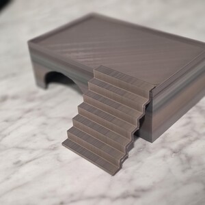 Puede incluir: Una estructura rectangular de varios niveles con un acabado liso de color gris oscuro. El objeto cuenta con una plataforma superior plana, un conjunto de escaleras integradas que conducen a la plataforma y una abertura arqueada hueca.