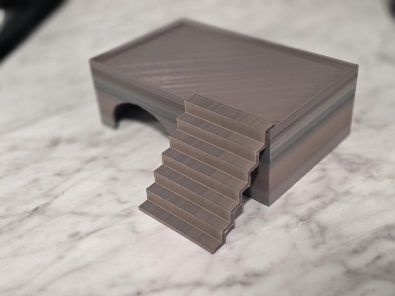Puede incluir: Una estructura rectangular de varios niveles con un acabado liso de color gris oscuro. El objeto cuenta con una plataforma superior plana, un conjunto de escaleras integradas que conducen a la plataforma y una abertura arqueada hueca.