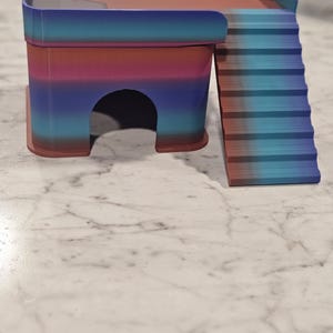 May include: A small, multi-level pet hideout featuring a gradient color scheme of blue, purple, and reddish-brown. The structure includes a rectangular platform with a protective railing, a ground-level arched entrance, and a set of stairs.
