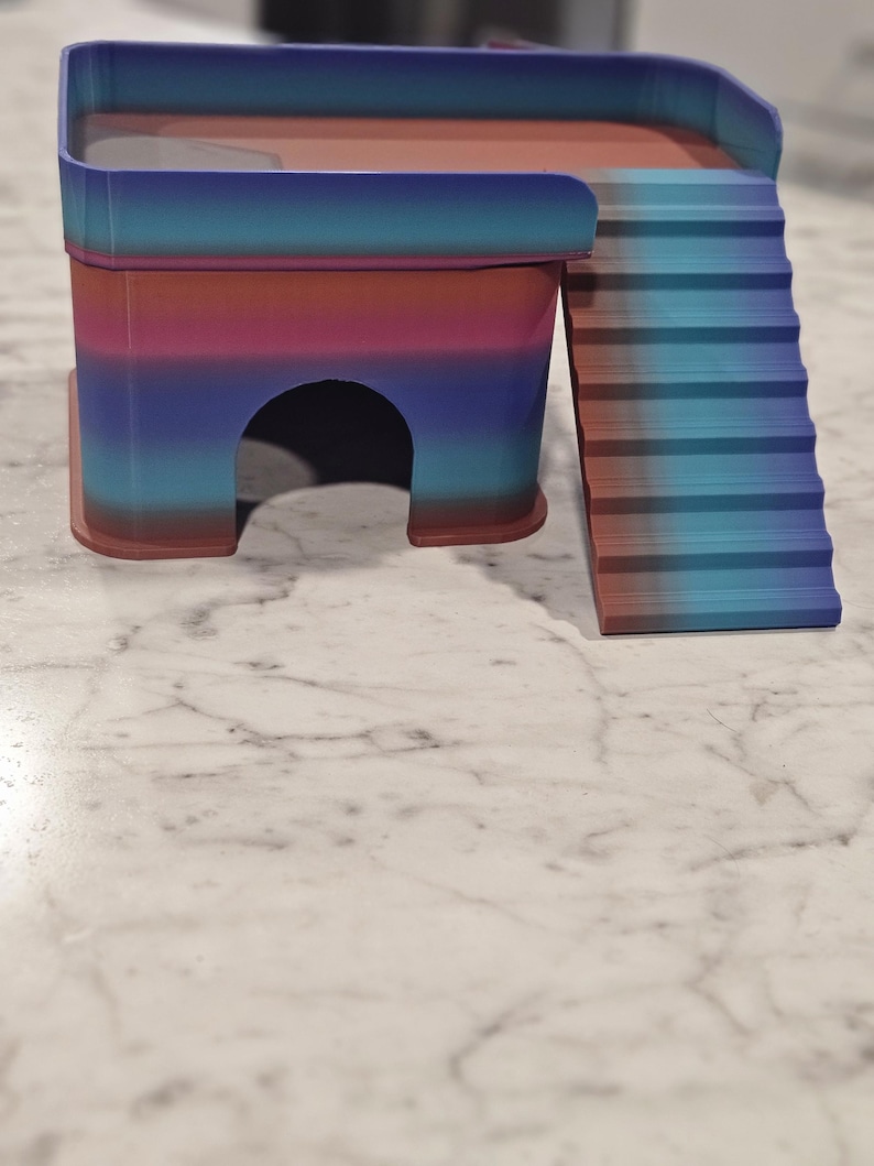 May include: A small, multi-level pet hideout featuring a gradient color scheme of blue, purple, and reddish-brown. The structure includes a rectangular platform with a protective railing, a ground-level arched entrance, and a set of stairs.