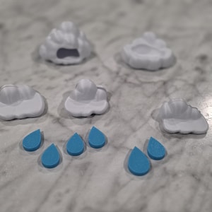 May include: A collection of five white cloud-shaped objects and five small blue teardrop-shaped pieces. The cloud shapes have rounded, puffy contours, while the blue pieces are flat, solid, and shaped like raindrops, arranged in a scattered formation.