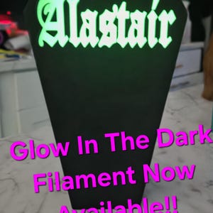 May include: A black coffin-shaped sign with the name "Alastair" in glowing green lettering. The sign also has the text "Glow In The Dark Filament Now Available!!" in pink.