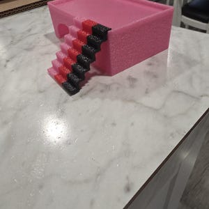Puede incluir: Una plataforma rectangular rosa con una abertura arqueada hueca en un lado y una escalera adjunta. La escalera presenta un patr&oacute;n de degradado de color que transiciona de rosa claro en la base a rojo en el medio y negro en el borde superior.