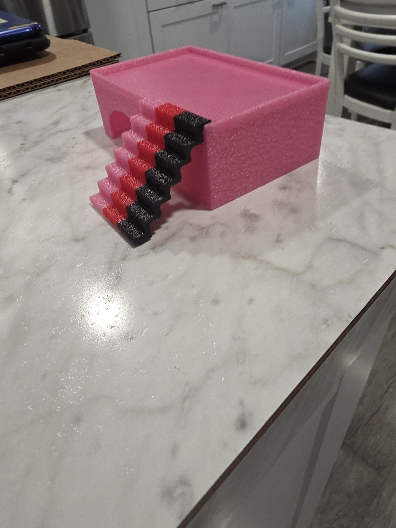 Puede incluir: Una plataforma rectangular rosa con una abertura arqueada hueca en un lado y una escalera adjunta. La escalera presenta un patr&oacute;n de degradado de color que transiciona de rosa claro en la base a rojo en el medio y negro en el borde superior.