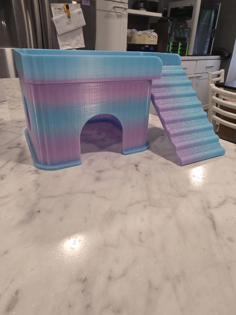 May include: A 3D printed pet hideout featuring a rectangular structure with a rounded arched entrance and an attached staircase leading to the top platform. The object displays a gradient color pattern transitioning between light blue and soft purple.
