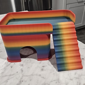 May include: A multi-colored, gradient-patterned small animal hideout featuring a rectangular platform with a curved railing, a tunnel-like opening at the base, and a set of stairs leading up to the top level, displaying a vibrant rainbow color scheme.