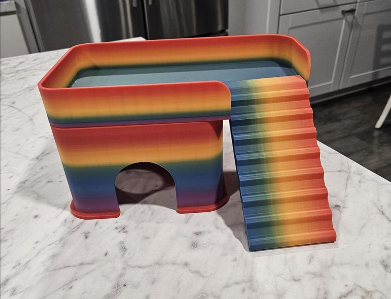 May include: A multi-colored, gradient-patterned small animal hideout featuring a rectangular platform with a curved railing, a tunnel-like opening at the base, and a set of stairs leading up to the top level, displaying a vibrant rainbow color scheme.