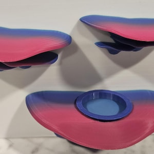 May include: A set of four wall-mounted shelves in a bi-color design. The shelves are a combination of blue and pink, with a rounded, cloud-like shape. One shelf has a small, circular blue dish.