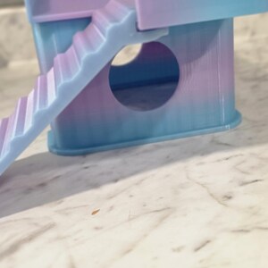 May include: A small, multi-level pet hideout featuring a gradient of pastel blue and purple colors. The structure includes a circular entrance hole on the front panel, a flat top platform, and a set of stairs leading up to the platform level.