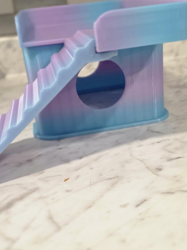 May include: A small, multi-level pet hideout featuring a gradient of pastel blue and purple colors. The structure includes a circular entrance hole on the front panel, a flat top platform, and a set of stairs leading up to the platform level.
