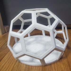 May include: A white, geometric, geodesic dome-shaped decorative object. The openwork design features interconnected pentagons and hexagons, creating a cage-like structure. The base is a solid white circle. The object is displayed on a wooden surface.