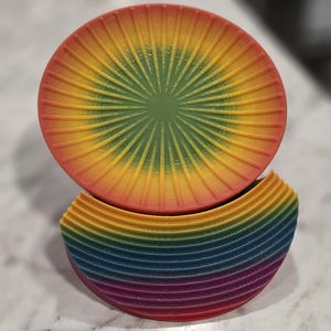 May include: A circular, 3D-printed object featuring a vibrant rainbow gradient. The top section displays a sunburst pattern radiating from the center, while the bottom section consists of stacked, curved layers in a spectrum of red, orange, yellow, green, blue, and purple.