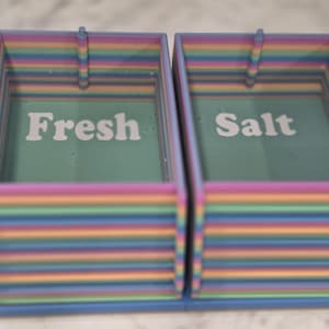 May include: Two rectangular storage containers with horizontal rainbow-colored stripes. The left container has the word Fresh printed in white on a teal base, and the right container has the word Salt printed in white on a teal base.