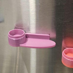 May include: Three magnetic shelves attached to a metallic surface. Each shelf features a circular holder and a flat, oval-shaped platform. The shelves are colored in light blue, light purple, and a combination of pink and coral orange.
