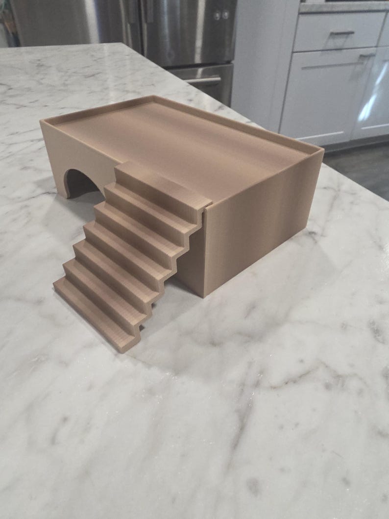 Puede incluir: Una plataforma rectangular de color beige con una superficie superior plana y un borde elevado. La estructura presenta una abertura arqueada en un lado y una escalera de seis pelda&ntilde;os que conduce al nivel de la plataforma, en material mate.