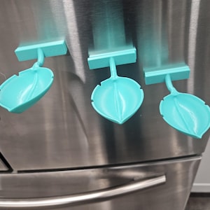 May include: Three turquoise leaf-shaped magnets are attached to a stainless steel refrigerator. Each magnet has a rectangular top piece and a curved stem connecting to the leaf shape. The magnets are evenly spaced and appear to be 3D-printed.