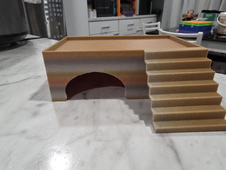 Puede incluir: Un escondite para mascotas de color tostado y multicolor que presenta una plataforma superior plana, una entrada en arco semicircular en la parte frontal y un conjunto de escaleras unidas al lado derecho que conducen al nivel superior.