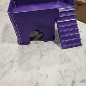 May include: A purple 3D-printed pet hideout featuring a raised platform with a surrounding wall and an attached staircase. The structure includes a hollow arched entrance at the base, providing a sheltered space for small animals to rest or climb.