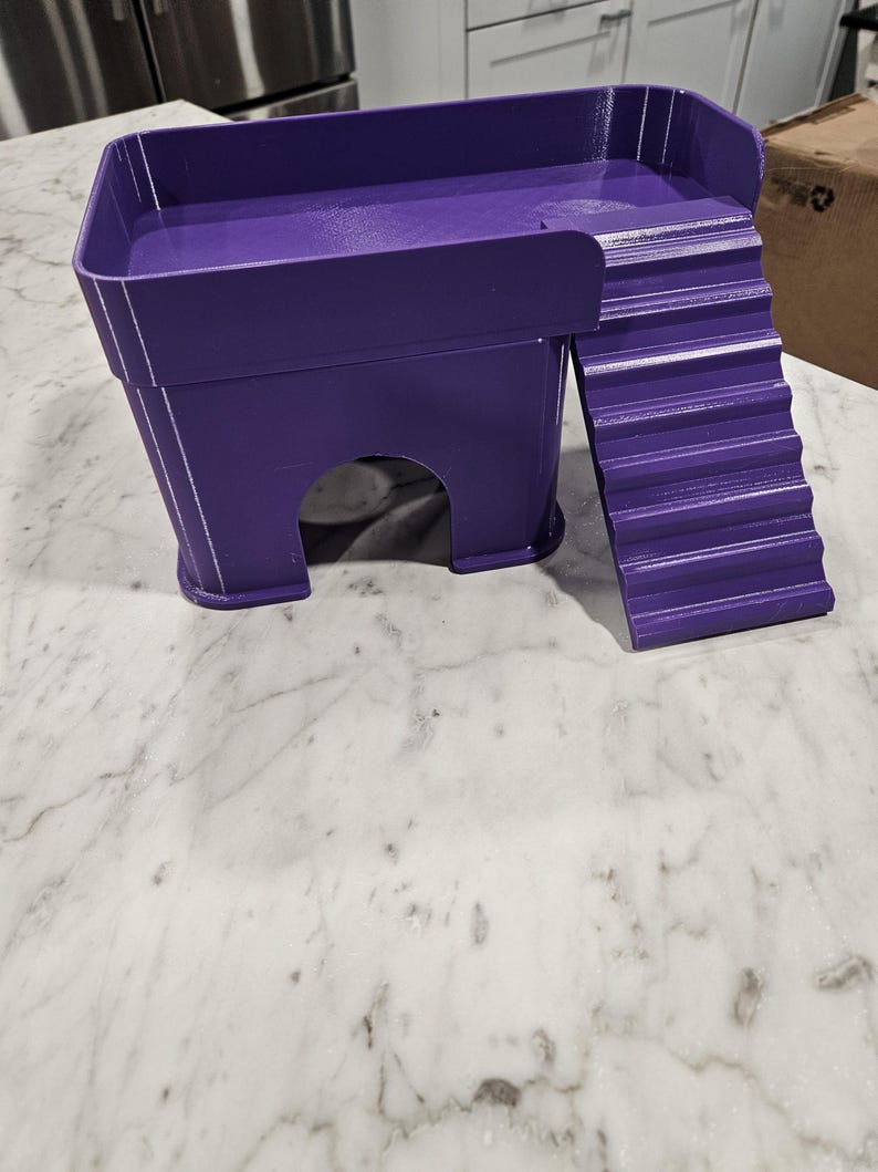 May include: A purple 3D-printed pet hideout featuring a raised platform with a surrounding wall and an attached staircase. The structure includes a hollow arched entrance at the base, providing a sheltered space for small animals to rest or climb.