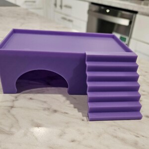 Puede incluir: Un escondite para mascotas de color morado s&oacute;lido que presenta una plataforma superior rectangular, una entrada en arco semicircular en la parte frontal y un conjunto de escaleras integradas que conducen al techo plano, hecho de material liso y mate.