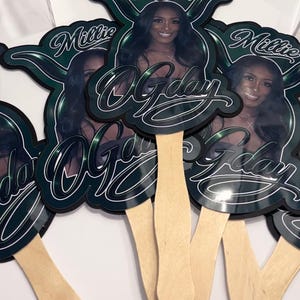 May include: A collection of decorative hand fans featuring a woman's image, the name "Millie", and the phrase "O'Gday" in a stylized font. The fans have a dark green and black color scheme with a wooden handle.
