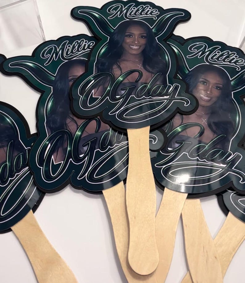 May include: A collection of decorative hand fans featuring a woman's image, the name "Millie", and the phrase "O'Gday" in a stylized font. The fans have a dark green and black color scheme with a wooden handle.