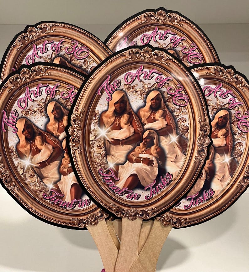 May include: A collection of oval-shaped hand fans. Each fan features a portrait of a person in a gold frame with the text "The Art of 30" in pink script. The fans have wooden handles.