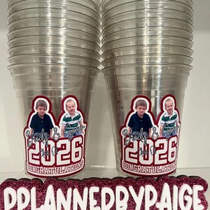 May include: Two stacks of clear plastic cups featuring a custom decal with photos of two children, the names Brody & Gavin, the text Class of 2026, and the word CONGRATULATIONS. A glittery pink sign in front displays PPLANNEDBYPAIGE with social media icons.