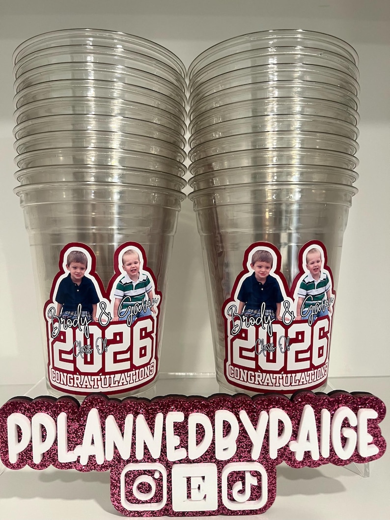 May include: Two stacks of clear plastic cups featuring a custom decal with photos of two children, the names Brody & Gavin, the text Class of 2026, and the word CONGRATULATIONS. A glittery pink sign in front displays PPLANNEDBYPAIGE with social media icons.