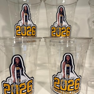 May include: Four clear plastic disposable cups featuring a custom decal. The decal displays a photograph of a graduate wearing a sash, the name Sydney, the year 2026 in large yellow font, and the text Class of 2026 Congratulations.