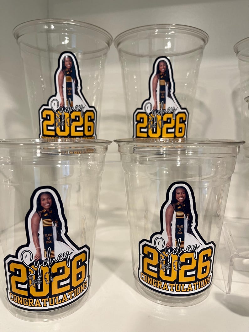May include: Four clear plastic disposable cups featuring a custom decal. The decal displays a photograph of a graduate wearing a sash, the name Sydney, the year 2026 in large yellow font, and the text Class of 2026 Congratulations.