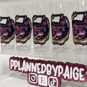Pu&ograve; includere: Cinque bicchieri alti trasparenti con un'etichetta grafica con il testo Welcome to Big Body's Trap, una persona e un'auto rosa. Sotto i bicchieri c'&egrave; un'insegna rosa scuro glitterata con testo bianco che recita PPLANNEDBYPAIGE.