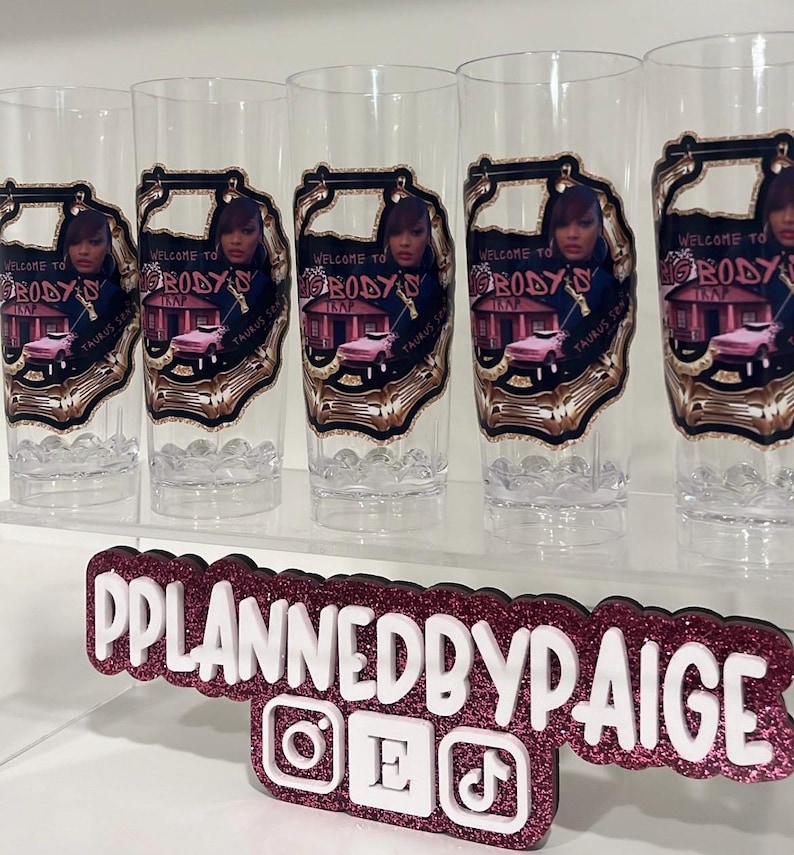 Pu&ograve; includere: Cinque bicchieri alti trasparenti con un'etichetta grafica con il testo Welcome to Big Body's Trap, una persona e un'auto rosa. Sotto i bicchieri c'&egrave; un'insegna rosa scuro glitterata con testo bianco che recita PPLANNEDBYPAIGE.