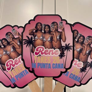 May include: Pink hand fans with a photo of three women in silver bikinis, palm trees, and the text "Rene Takes 26 in Punta Cana." The fans have black borders and wooden handles.