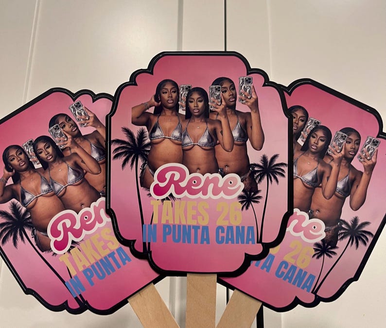 May include: Pink hand fans with a photo of three women in silver bikinis, palm trees, and the text "Rene Takes 26 in Punta Cana." The fans have black borders and wooden handles.