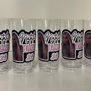 Pu&ograve; includere: Cinque bicchieri in plastica trasparente con un adesivo personalizzato raffigurante il ritratto fotografico di una persona con lunghi capelli biondi. Il testo Natalie Takes 26 &egrave; in un carattere nero e rosa con contorno bianco su sfondo nero.