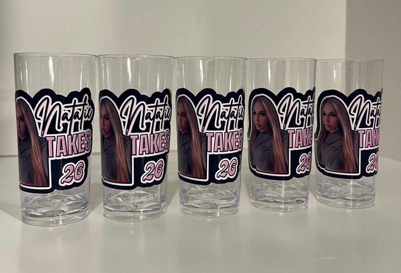 Pu&ograve; includere: Cinque bicchieri in plastica trasparente con un adesivo personalizzato raffigurante il ritratto fotografico di una persona con lunghi capelli biondi. Il testo Natalie Takes 26 &egrave; in un carattere nero e rosa con contorno bianco su sfondo nero.