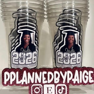 May include: Two clear plastic cups featuring a graphic of a graduate in a cap and gown with the text Jada, Class of 2026, and Congratulations. In front is a glittery pink and white sign reading PPLANNEDBYPAIGE with Instagram, Etsy, and TikTok icons.