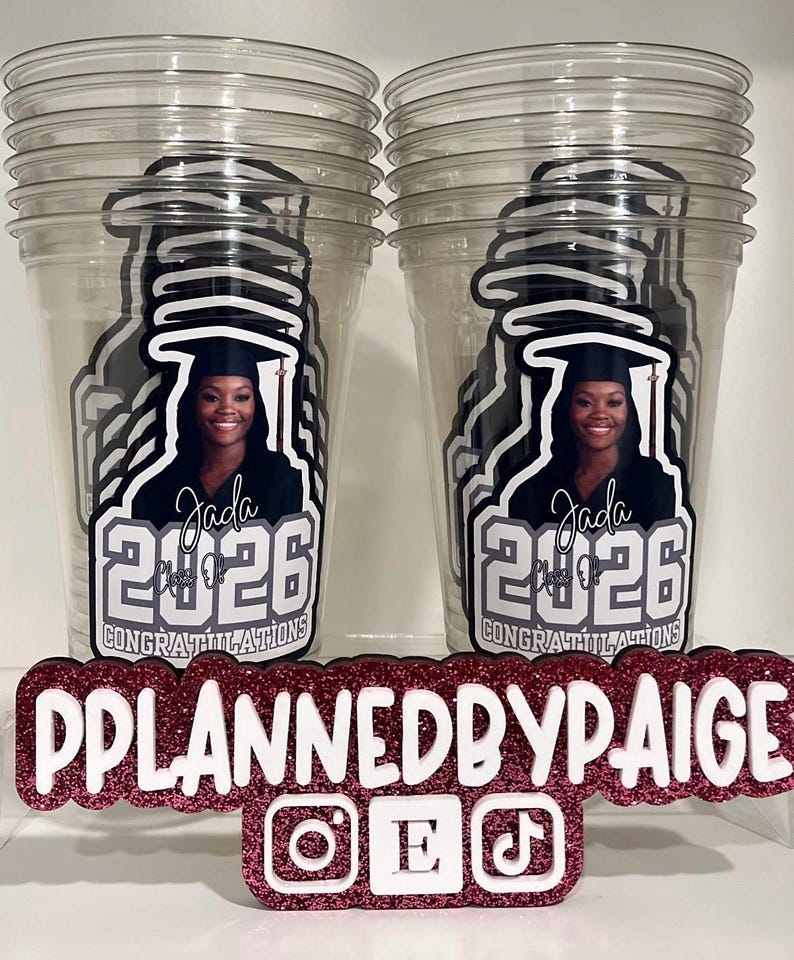 May include: Two clear plastic cups featuring a graphic of a graduate in a cap and gown with the text Jada, Class of 2026, and Congratulations. In front is a glittery pink and white sign reading PPLANNEDBYPAIGE with Instagram, Etsy, and TikTok icons.