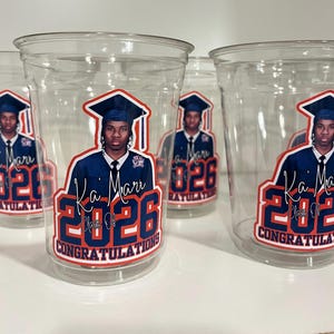 May include: Clear plastic disposable cups featuring a graduation-themed graphic. The design includes a portrait of a graduate in a cap and gown, the name Ka'Mari, the text Class of 2026, and the word CONGRATULATIONS in bold blue and red lettering.