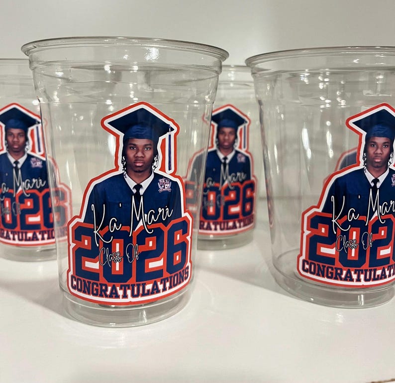 May include: Clear plastic disposable cups featuring a graduation-themed graphic. The design includes a portrait of a graduate in a cap and gown, the name Ka'Mari, the text Class of 2026, and the word CONGRATULATIONS in bold blue and red lettering.