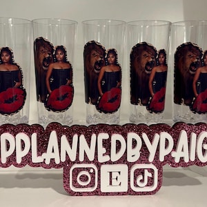 Pu&ograve; includere: Cinque bicchieri trasparenti con una stampa grafica di una persona in top nero accanto a un leone ruggente e un design di labbra rosse. Un cartello rosa glitterato mostra il testo PPLANNEDBYPAIGE con icone dei social media.