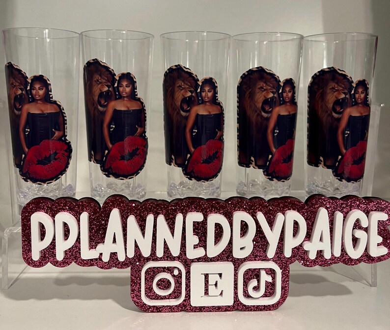 Pu&ograve; includere: Cinque bicchieri trasparenti con una stampa grafica di una persona in top nero accanto a un leone ruggente e un design di labbra rosse. Un cartello rosa glitterato mostra il testo PPLANNEDBYPAIGE con icone dei social media.