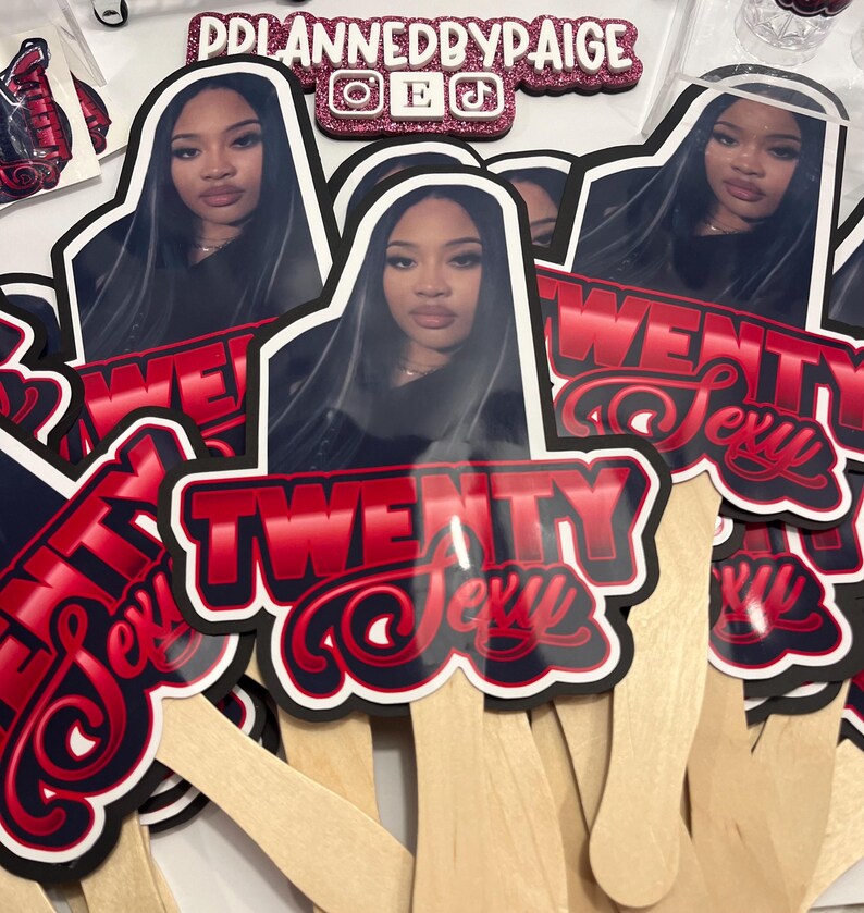 May include: A collection of fan-shaped party favors. Each fan features a photo of a woman and the words "TWENTY Sexy" in red and white lettering. The fans are mounted on wooden sticks. The top of the image shows a glittery sign that says "DDPLANNEDBYPAIGE".