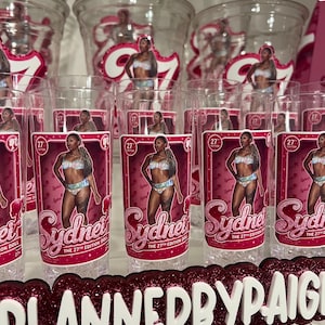 Pu&ograve; includere: Una collezione di bicchieri in plastica trasparente con un'etichetta rettangolare rosa raffigurante una bambola in un completo a due pezzi. L'etichetta include il testo Sydney, The 27th Edition Doll e un piccolo logo. Un'insegna glitterata riporta PLANNEDBYPAIGE.
