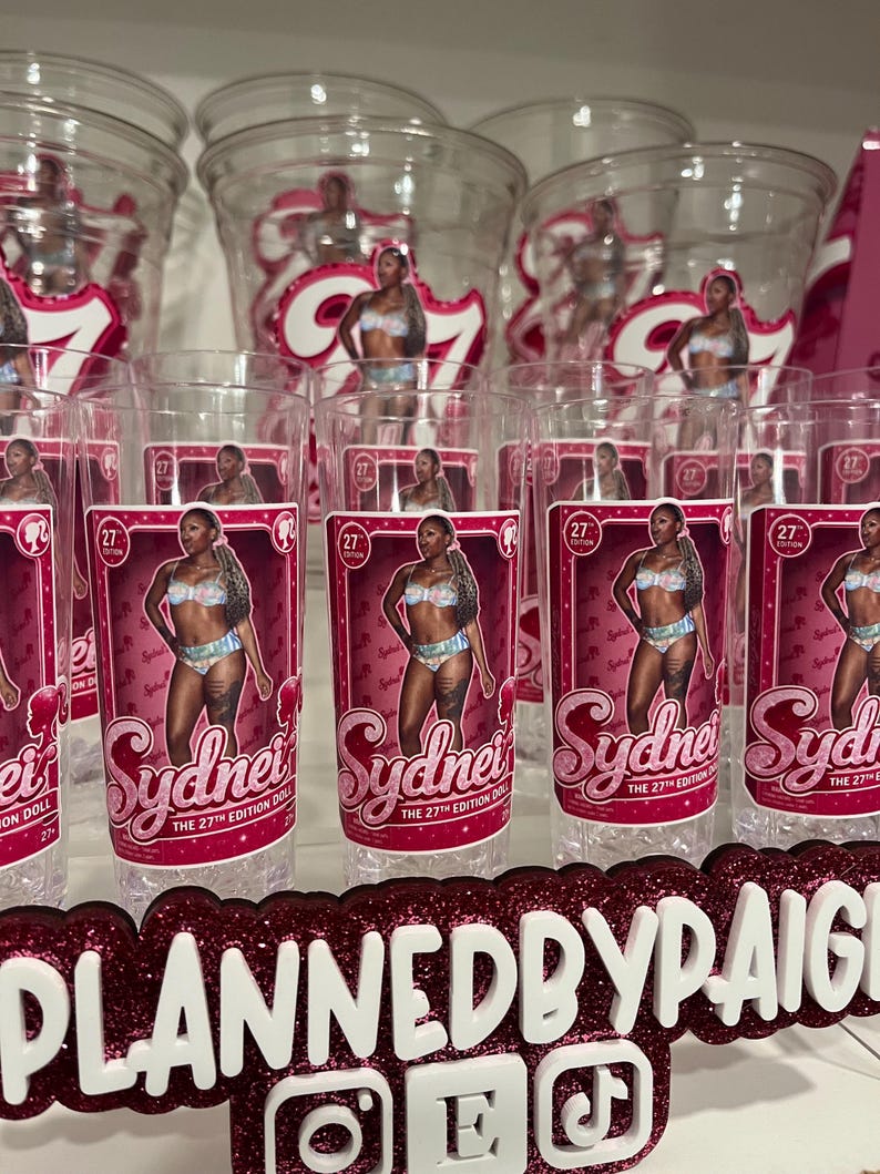 Pu&ograve; includere: Una collezione di bicchieri in plastica trasparente con un'etichetta rettangolare rosa raffigurante una bambola in un completo a due pezzi. L'etichetta include il testo Sydney, The 27th Edition Doll e un piccolo logo. Un'insegna glitterata riporta PLANNEDBYPAIGE.