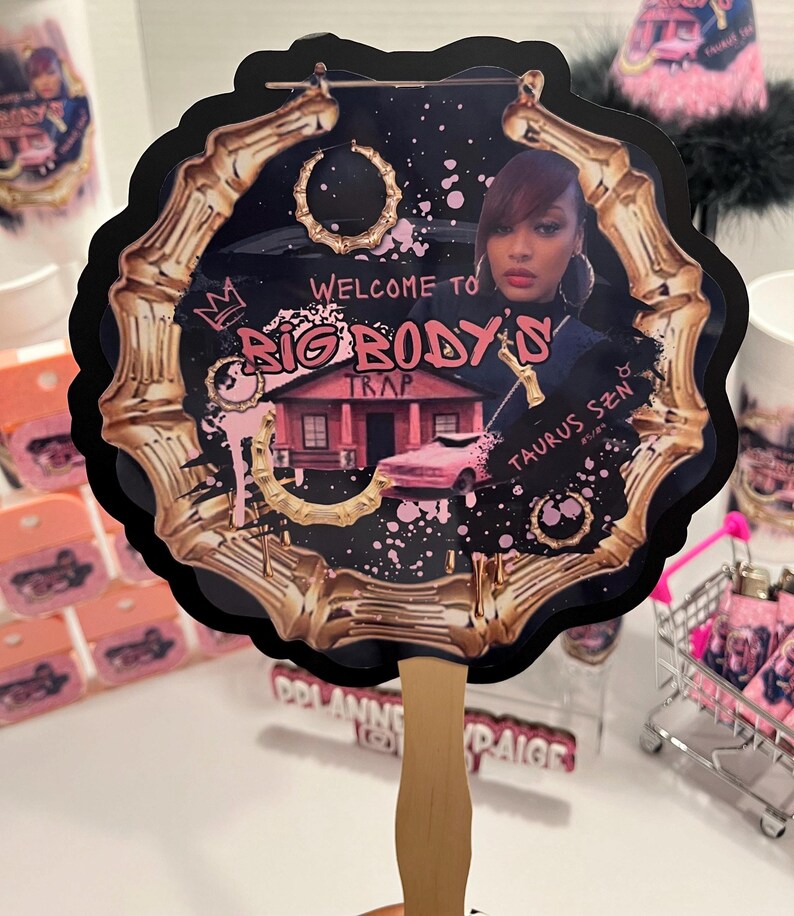 May include: A decorative fan with a black scalloped edge and a bamboo-style gold frame. The fan features a woman's image, text that reads "Welcome to Big Body's Trap", and pink accents. The handle is made of light wood.