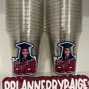May include: Two clear plastic cups featuring a graduation-themed decal with a photo of a person in a cap and gown, the name Kylah Symone, and the text Class of 2026 Congratulations. A glittery sign in front displays PPLANNEDBYPAIGE with social media icons.