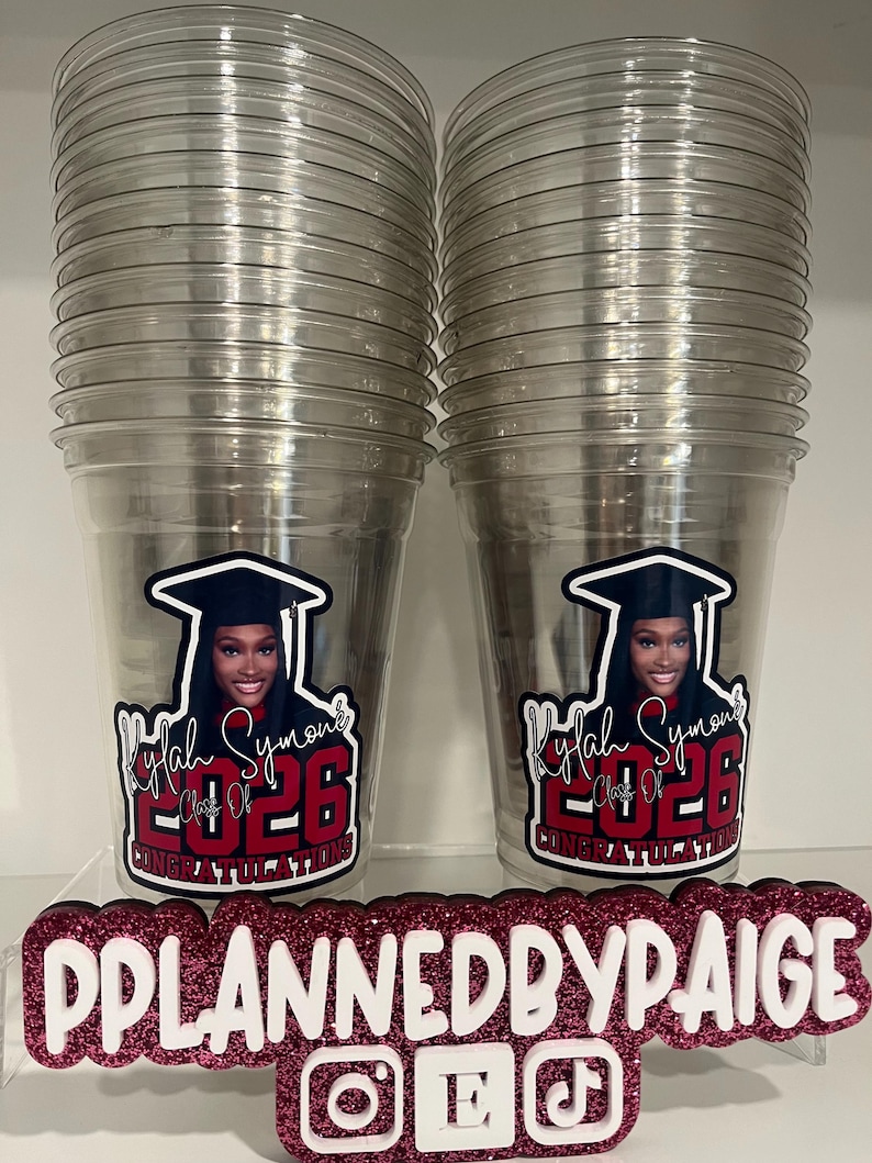 May include: Two clear plastic cups featuring a graduation-themed decal with a photo of a person in a cap and gown, the name Kylah Symone, and the text Class of 2026 Congratulations. A glittery sign in front displays PPLANNEDBYPAIGE with social media icons.