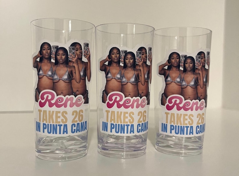 Pu&ograve; includere: Tre bicchieri in plastica trasparente con un adesivo fotografico di una persona in bikini argento che tiene in mano un telefono. Il testo recita "Rene TAKES 26 IN PUNTA CANA" in lettere rosa, oro e blu. Il design &egrave; ripetuto su ogni bicchiere.